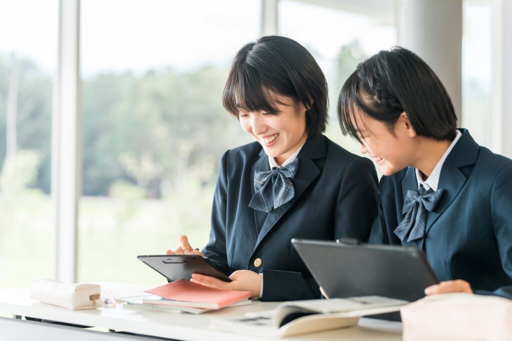 Two female students in school uniforms are sitting together at a table, smiling and looking at a tablet. There are notebooks and stationery on the table near them, with a bright and airy background.